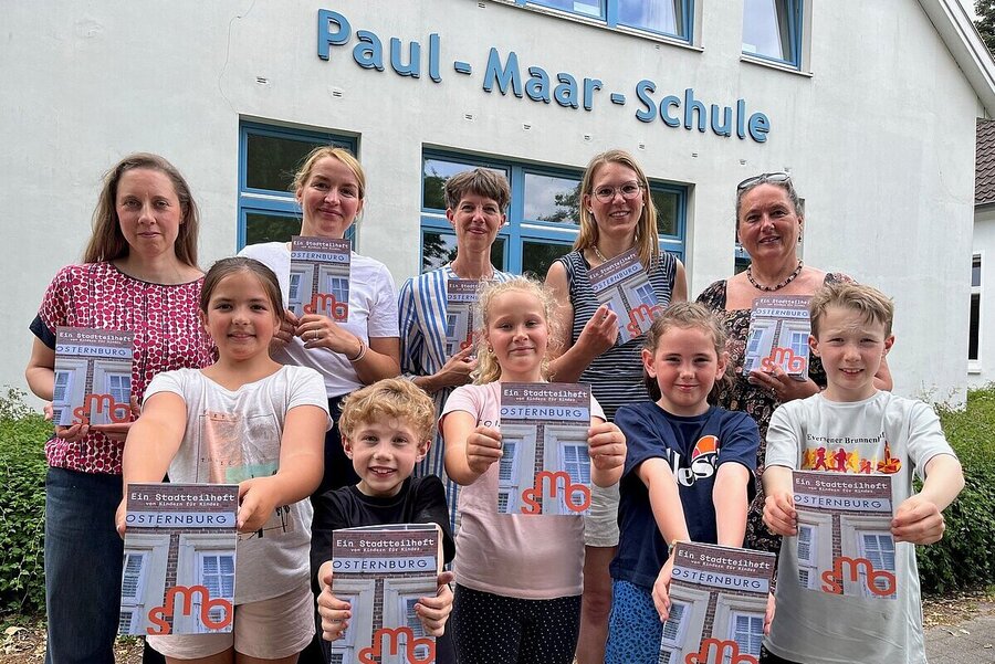 Präsentation des Stadtteilhefts an der Grundschule Paul-Maar-Schule, von links: Laura Keppel und Carolin Werel (Konzept und Gestaltung), Sandrine Teuber (Bildung und Vermittlung städtische Museen), Gesa Neugebauer (Schulleitung), Geraldine Dudek (Verein Lebendiges Museum) mit fünf Kindern der Paul-Maar-Schule, Foto: Stadtmuseum Oldenburg