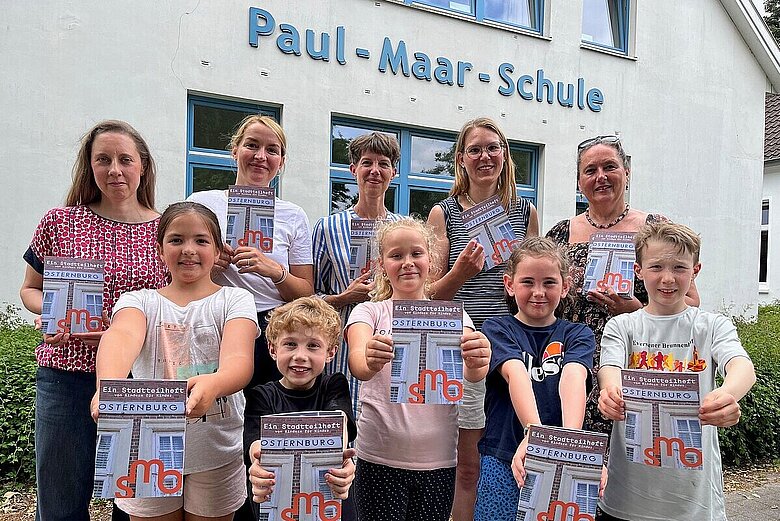 Präsentation des Stadtteilhefts an der Grundschule Paul-Maar-Schule, von links: Laura Keppel und Carolin Werel (Konzept und Gestaltung), Sandrine Teuber (Bildung und Vermittlung städtische Museen), Gesa Neugebauer (Schulleitung), Geraldine Dudek (Verein Lebendiges Museum) mit fünf Kindern der Paul-Maar-Schule, Foto: Stadtmuseum Oldenburg